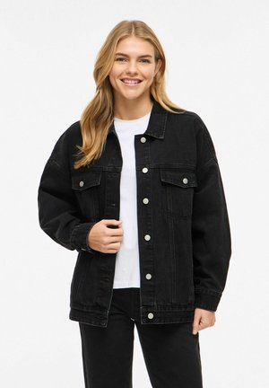 Jonge vrouw lacht, draagt een oversized zwarte spijkerjasje over een wit shirt en zwarte broek, staand voor een effen achtergrond.