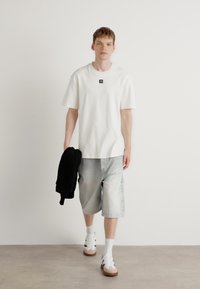 T-shirt bianca in cotone con un piccolo logo, abbinata a pantaloni corti in denim blu chiaro. Il modello indossa sneakers bianche e tiene un felpa nera.