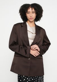 Brūns piemērots žakete ar vienu pogu, lieliem atlokiem un divām priekšējām kabatām, valkāts virs gaiši bēša satīna topa, uz vienkārša fona.