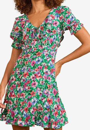 Kvinde iført grøn blomsterkjole med korte ærmer, rynket halsudskæring og kant, stående med den ene hånd på hoften.