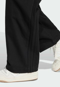 Svarta vida byxor i mjukt material med sidostänger, parat med vita sneakers med texturerade detaljer och en ljusbrun sula.