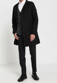 Svart trenchcoat med dubbelknäppt design, axelklaffar, över en grå tröja och rutiga byxor, ihop med svarta sneakers.