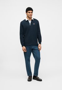 Άνδρας που στέκεται φορώντας μπλε μακρυμάνικο πόλο με λευκό γιακά, μπλε τζιν και μαύρα παπούτσια σε φόντο με απλό λευκό χρώμα.