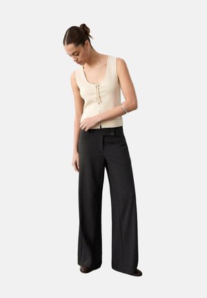 Donna che indossa un top beige senza maniche con dettaglio anteriore con lacci e pantaloni neri a gamba larga, guarda verso il basso con la mano vicino alla vita.