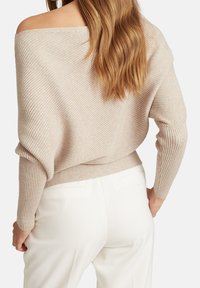 Pull beige à épaules dénudées avec une texture côtelée, manches longues et taille ajustée, associé à un pantalon blanc sur mesure.