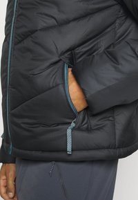 Giubbotto imbottito nero con un motivo trapuntato, dotato di cerniere e dettagli in teal. Include una tasca laterale con chiusura a cerniera e una consistenza morbida.