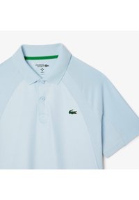 Ljusblå pikétröja med korta ärmar, texturerade sidopaneler, treknappars placket och grön accent på kragen. Har en grön krokodil-logotyp.