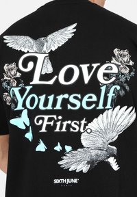 Zwarte t-shirt met witte en lichtblauwe graphics van vogels, vlinders, rozen en de tekst "Love Yourself First" in vette letters.