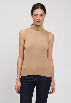 Donna con i capelli scuri raccolti in una coda di cavallo indossa un top a collo alto senza maniche color beige e jeans scuri, in piedi contro uno sfondo chiaro e uniforme.