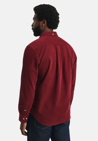 Man met kort, krullend haar en een baard, gekleed in een bordeauxrode blouse met lange mouwen en een donkere spijkerbroek, met zijn rug naar de camera gekeerd.