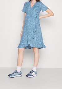 Blaues Wickelkleid mit kurzen Ärmeln, weißem Blumendruck und gerafftem Saum. Kombiniert mit blauen und marineblauen Turnschuhen und weißen Socken.