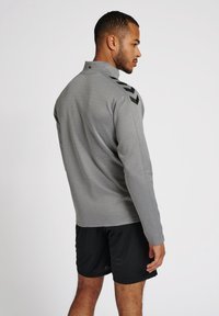 Pull-over gris à manches longues en tissu lisse, avec des détails en chevron noirs sur les épaules, associé à un short de sport noir.