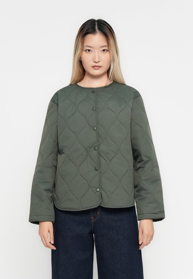 Femme portant un manteau matelassé olive en boutons et un pantalon en jean large bleu marine, se tenant devant un fond blanc.