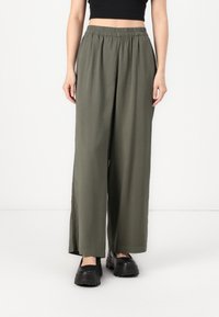 Un pantalon large en tissu vert olive avec une taille élastique, présentant des fentes sur les côtés et une texture lisse. Associé à des chaussures à plateforme noires.