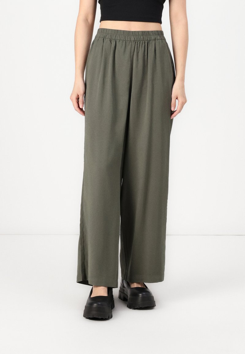 Un pantalon large en tissu vert olive avec une taille élastique, présentant des fentes sur les côtés et une texture lisse. Associé à des chaussures à plateforme noires.