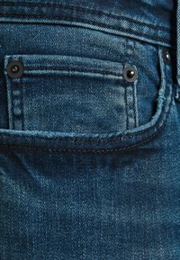 Dark blue denim jeans with a visible back pocket, featuring black button accents and contrasting stitching details. Textured fabric with a slight sheen.