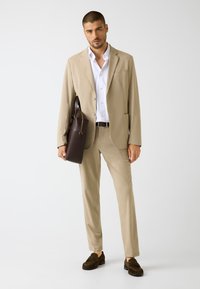 Completo beige su misura con giacca monopetto, pantaloni abbinati, camicia bianca e borsa in pelle marrone scuro. Mocassini in camoscio marrone.