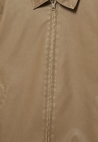 Detailní záběr přední části béžové kožené bundy, zobrazující límec, zapínání na zip a částečný pohled na kapsu na strukturovaném materiálu.