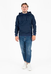 Felpa blu navy con un grande logo in rilievo, cappuccio con coulisse, tasca frontale e polsini a coste; abbinata a jeans azzurri e sneakers bianche.