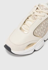 Shoe atlético de cor creme com parte superior em camurça e sintético, detalhes em dourado, design texturizado e sola de borracha preta. Apresenta detalhe do logótipo.