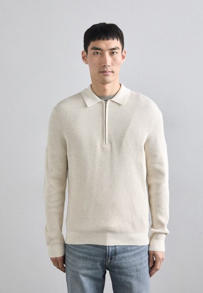 Pull en maille beige clair avec un col zippé, présentant une texture côtelée et des manches longues, associé à un jean bleu.