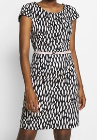 Femme portant une robe mi-longue à motifs noirs et blancs, manches courtes, avec une ceinture rose clair à la taille.