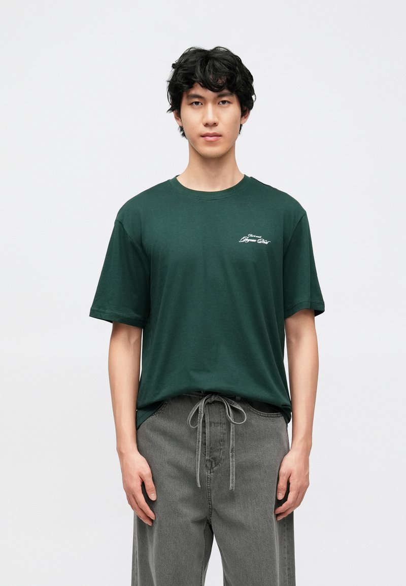 Camiseta de algodón verde con mangas cortas y cuello redondo, con un pequeño detalle de logo blanco en el pecho izquierdo. Combinada con pantalones grises.