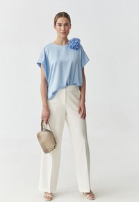 Blusa di raso azzurro chiaro a maniche corte con un accento floreale; abbinata a pantaloni bianchi a gamba larga e alta vita e a una borsa beige.