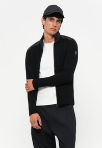 Μαύρο φούτερ με φερμουάρ και μακριά μανίκια, με ριμπ περίγραμμα και διακοσμητικά στις ώμους από υφή. Φορεμένο πάνω από λευκό πουκάμισο, με κομψό σχέδιο.