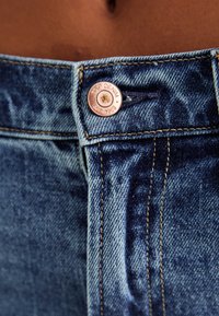 Blue denim jeans featuring a copper button with "GAP DENIM EST. 1969" engraved, visible stitching, and a classic five-pocket design.