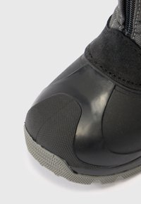 Nærbilde av en svart boots gummitåhette, teksturert såle og semsket overdel med glidelåsdetalj på en hvit bakgrunn.