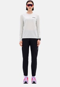 Hellgraues langärmeliges sportliches Oberteil mit schwarzem Logo, kombiniert mit schwarzen taillierten Hosen mit Reißverschlüssen und rosa Sportschuhen.