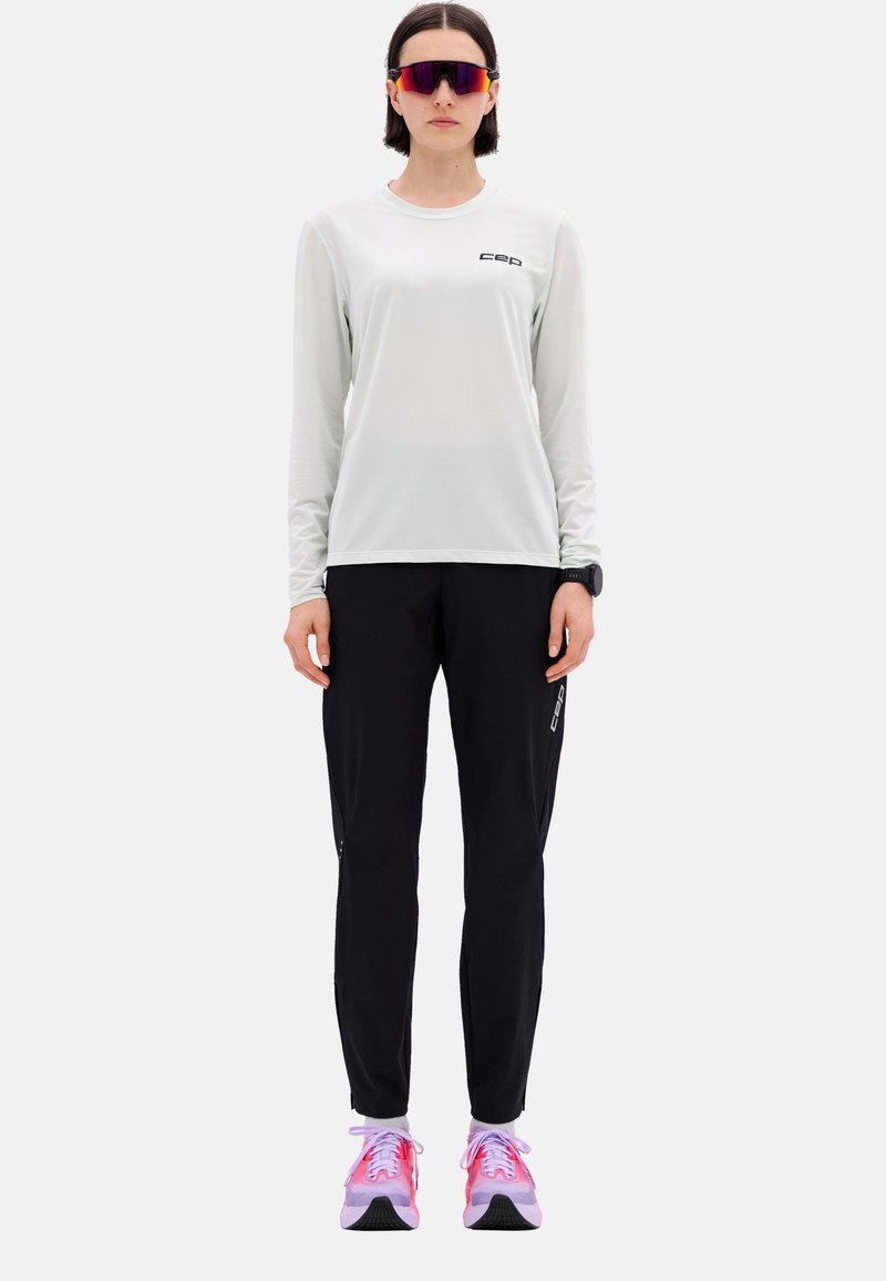 Hellgraues langärmeliges sportliches Oberteil mit schwarzem Logo, kombiniert mit schwarzen taillierten Hosen mit Reißverschlüssen und rosa Sportschuhen.