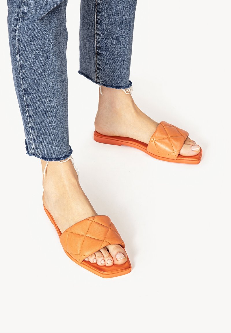 Sandales slides matelassées orange avec un bout carré, une semelle plate et une texture en cuir lisse, associées à un jean en denim effiloché.