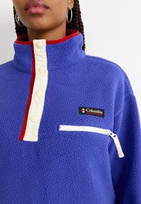 Pullover in pile di un vivace colore viola con colletto alto, dettagli rossi, bottoni a pressione e tasca sul petto con zip. Logo Columbia visibile.