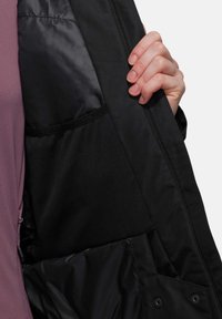 Schwarze Jacke innen mit glatter Satintextur und Netzfach. Eine Hand hält die Jacke offen und zeigt kontrastierende Stofflagen.