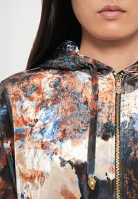 Mīksta, teksturēta auduma kapuce, izgatavota zilā, oranžā un pelēkā krāsā; komplektā ar zelta rāvējslodzi un dekoratīvu pogu.