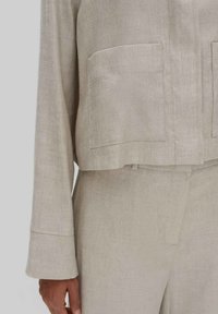Lichtbeige linnen jas met een cropped ontwerp, twee voorkwasten en lange mouwen. De stof heeft een gestructureerde afwerking.