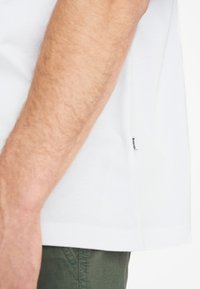 Camiseta blanca de algodón con mangas cortas, bordes rematados y una pequeña etiqueta de tela en un lado. La piel texturizada es visible en el brazo.