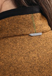 Fleecebekleidung aus einem strukturierten orange-braunen Stoff, mit schwarzem Kragen und kontrastierenden grünen Nähten. Beinhaltet ein kleines graues Stoffdetail.