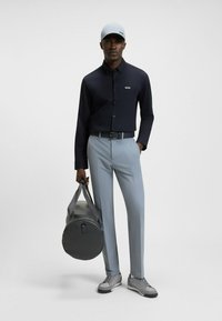 Navy long-sleeve shirt, light blue trousers, gray sneakers, and matching cap. Holds a gray duffel bag. Simple, modern design with logos.