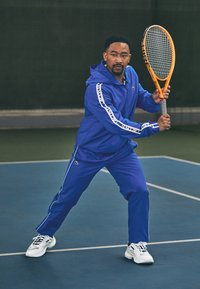 Kék sportos melegítő, fehér díszítéssel, zipzáros kapucnis pulóverrel és elasztikus derékpánttal. Egy férfi narancssárga teniszütőt tart a pályán.