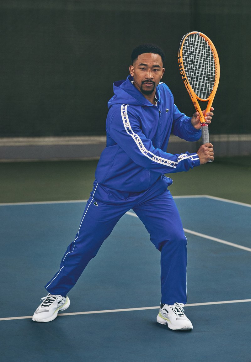 Kék sportos melegítő, fehér díszítéssel, zipzáros kapucnis pulóverrel és elasztikus derékpánttal. Egy férfi narancssárga teniszütőt tart a pályán.