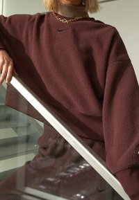 Vinröd fleecetröja med hög krage, med en svart Nike-logotyp och ett halsband i guldkedja. Modellen lutar sig mot ett glasräcke.