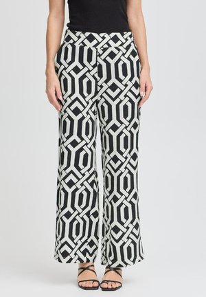 Pantalons ample à motif géométrique noir et blanc, avec une taille haute et un tissu lisse. Associés à des sandales noires à lanières.