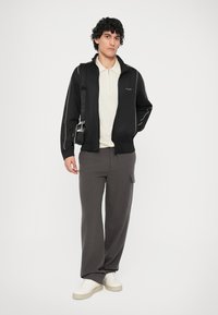 Chaqueta negra con cremallera y ribete blanco, camiseta polo beige, pantalones de chándal grises y zapatillas blancas. El modelo está de pie con un bolso bandolera.