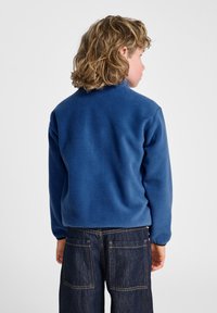 Blauwe fleecejas met een hoge kraag en lange mouwen, gemaakt van zacht materiaal. Gecombineerd met donkere spijkerbroek met twee achterzakken.