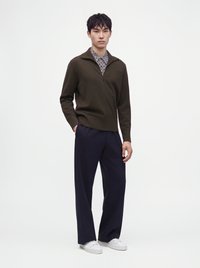 Homme portant un pull zippé vert foncé sur une chemise à motifs, pantalon large bleu marine et baskets blanches, debout sur fond blanc.