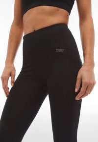 Leggings neri a vita alta realizzati in tessuto elasticizzato. Caratterizzati da una tasca laterale e un dettaglio logo discreto sulla cintura. Texture liscia.