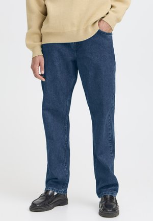 Person iført mørkeblå jeans med lige ben, beige sweater og sorte lædersko, stående med den ene hånd i lommen på hvid baggrund.